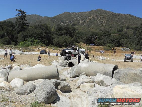 Modified off-road truck with large tires on rocks, other vehicles and people in background
