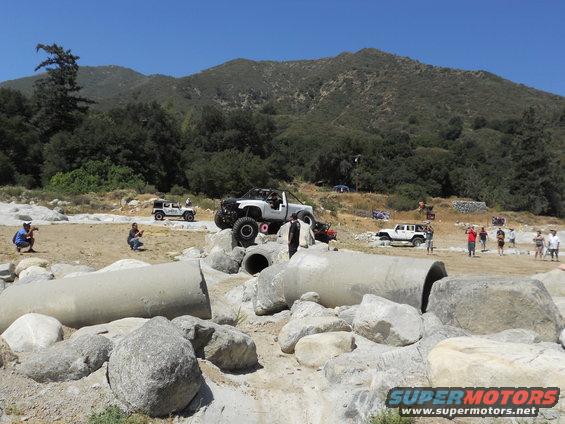 Off-road vehicles on a rocky course with large concrete pipes and hills in background