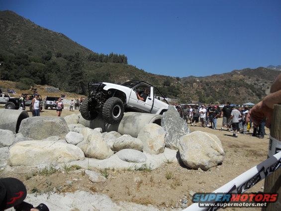 White off-road vehicle with large tires climbing over rocks, crowd watching