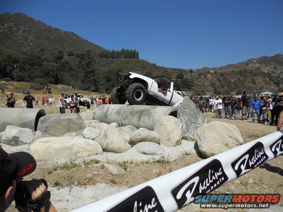 White off-road truck climbing over concrete pipes with crowd watching. Another white SUV is visible in the background