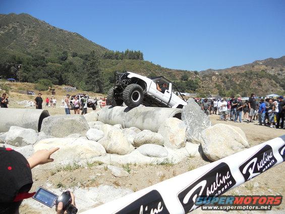 White off-road truck with a roll cage on large rocks, driver visible