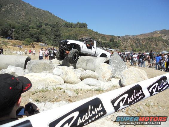 Modified white off-road truck climbing over concrete pipes and rocks. Spectators watch in the background