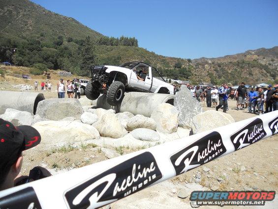 White off-road truck climbing over large rocks and concrete pipes with spectators watching. Raceline Wheels banner in foreground