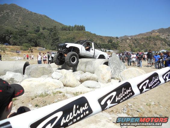 White off-road truck with large tires climbing over concrete pipes and rocks, with spectators watching