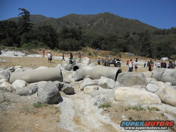 Off-roaders gathered around a white Jeep at a rocky obstacle course with large concrete pipes