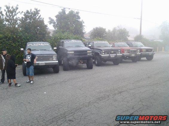 Several lifted pickup trucks parked in a row, some with text on the windshields. People stand near the first truck