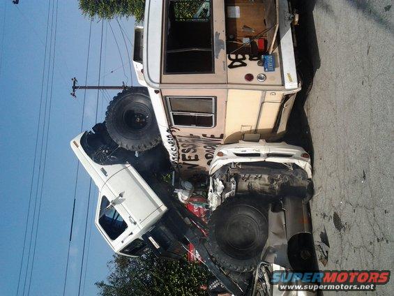 Wrecked white pickup truck on its side, partially under a damaged RV. Another white car is crushed between them
