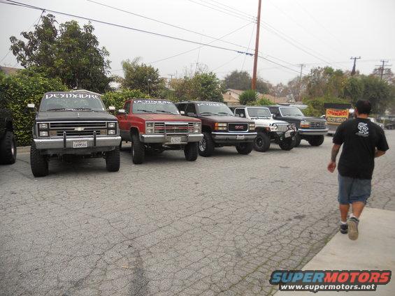 Several lifted trucks and SUVs parked in a lot, including a black Chevy, a red Chevy, a maroon GMC, a white Jeep, and a black Ford. A person walks away from the camera