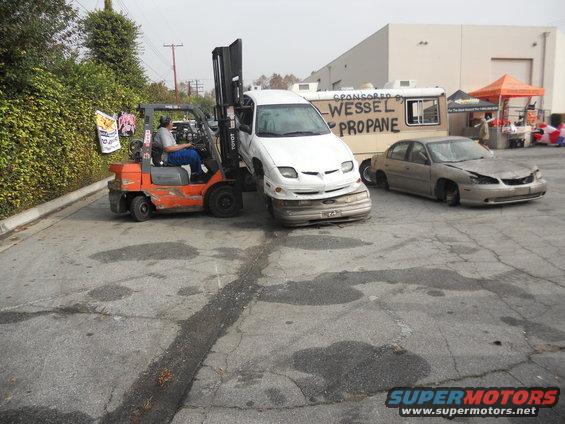 Forklift lifting a white car with a damaged front end; another damaged car sits nearby. "WESSEL PROPANE" is written on a trailer