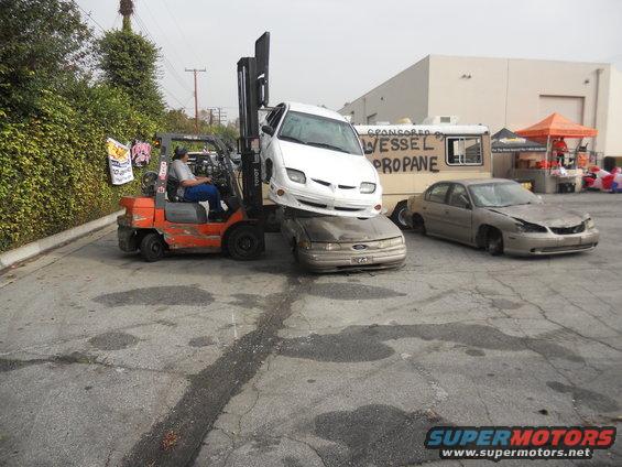 forklift lifting a white car onto a gold car, another gold car sits nearby