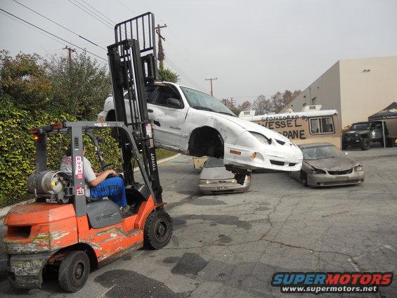 Forklift lifting a white car, with two other cars below it. A sign on a trailer reads 'SPONSORED BY VESSEL LOAN'