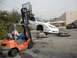 1986 Ford Bronco - A forklift is lifting a white car, which is suspended over two other vehicles in a yard.