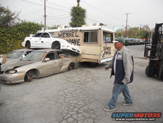 Three wrecked cars are stacked and leaning against a trailer. A man walks past