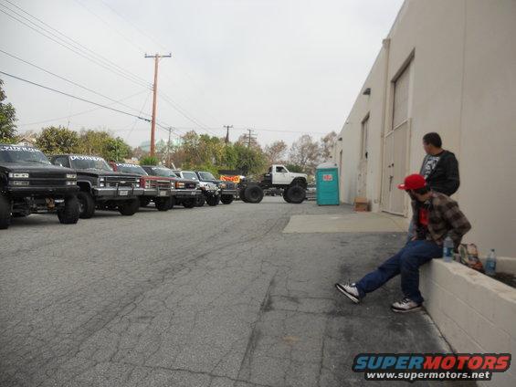 Several lifted pickup trucks parked in a lot, with two guys hanging out by a building