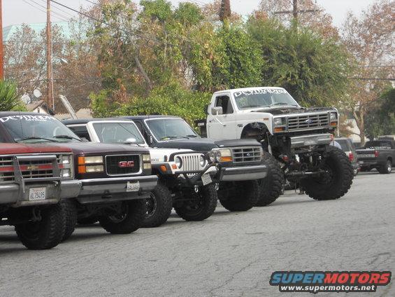 White Ford F-Series truck with massive lift kit and tires, parked in a line with other modified vehicles