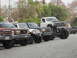 1986 Ford Bronco - Several modified trucks and SUVs are parked in a line, with a heavily lifted white Ford F-Series truck prominently displayed.