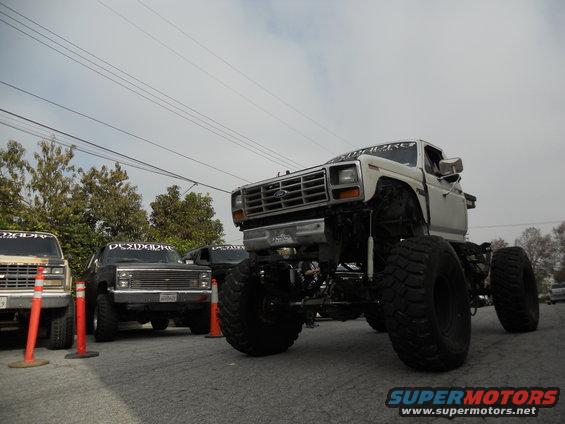 Lifted white Ford truck with large tires, front 3/4 view. Other trucks visible