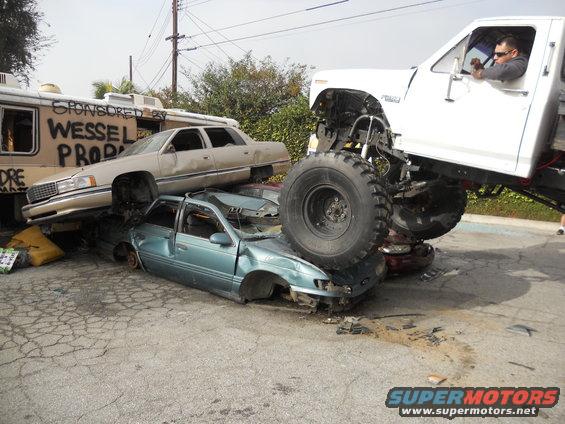 Monster truck on top of crushed cars, one car is on top of another. A trailer is behind them with graffiti