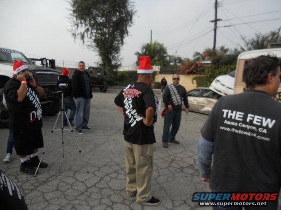 People in Santa hats stand near damaged cars, one with a white front end visible