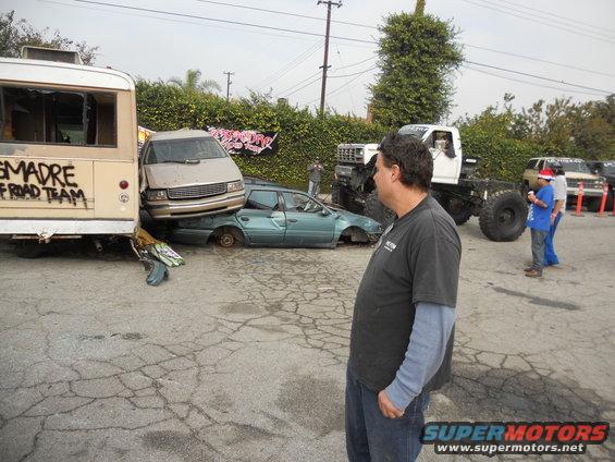 Crushed cars and a bus are piled up next to a monster truck. A man stands looking at the scene