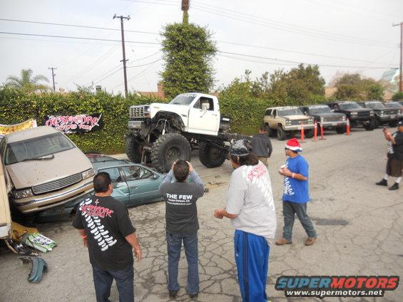 White lifted truck towering over a crushed beige sedan and a teal sedan, with several other trucks parked in the background