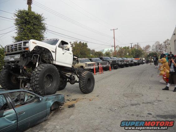 White monster truck with large tires positioned over a crushed teal sedan. A line of trucks is parked in the background