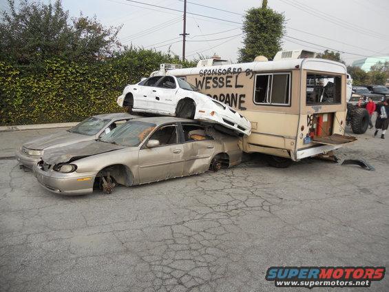 Damaged cars piled up, one on top of another, next to a bus with text on its side