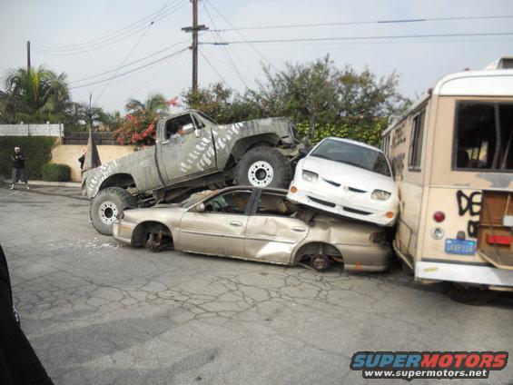 Monster truck on top of a sedan, which is on top of another sedan, next to a bus. Cars are wrecked