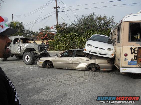lifted pickup truck, a crushed sedan, a white car on top of the sedan, and a bus are in a junkyard scene