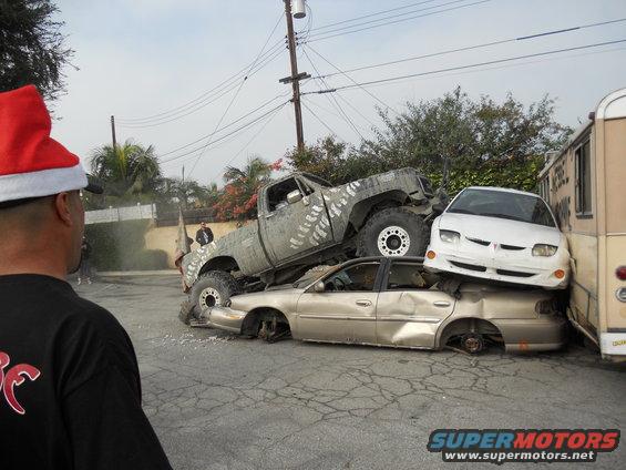 Crushed cars with a lifted truck on top, a white car on the side, and a bus
