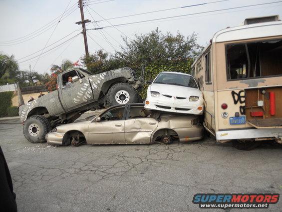 Lifted pickup truck crushing a sedan, with a white car on top and a bus to the right