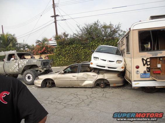 Wrecked gold sedan crushed by white car, which is next to a bus. A large truck is in front of the sedan. All vehicles appear damaged