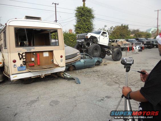Monster truck on top of a crushed car, with a bus to the left. Someone is filming