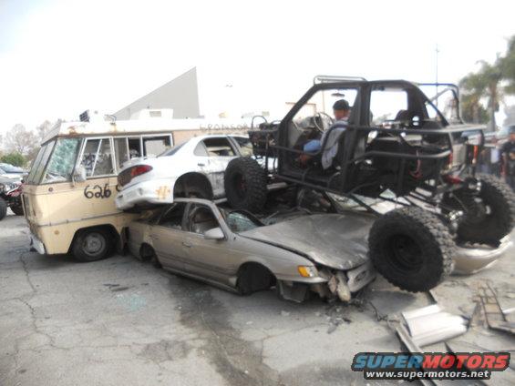 Crushed cars and a van under a custom buggy, with a driver visible