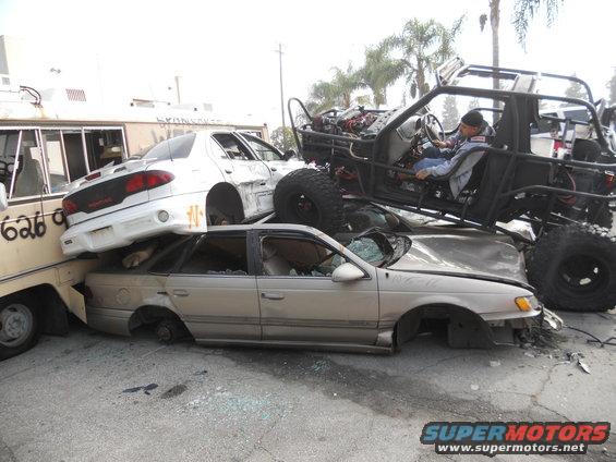 Several wrecked cars are piled up, with a custom off-road vehicle on top. A person is in the driver's seat of the off-road vehicle