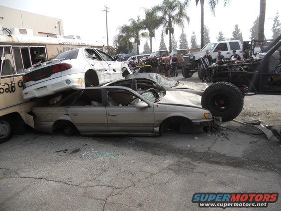 Several wrecked cars piled up, one on top of another, with a large off-road vehicle in the background