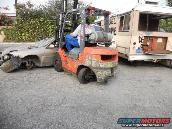 Damaged sedan being lifted by an orange Toyota forklift, with a beige bus in the background. Text on bus reads 'DIESEL PANE'