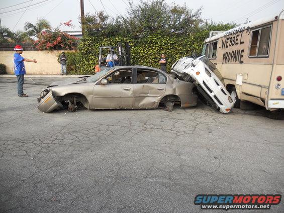 Damaged sedan crushed by a truck, with a forklift and people nearby