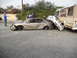 1986 Ford Bronco - A sedan is severely damaged, crushed by a truck, with a forklift and people present at the scene.