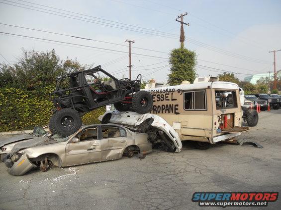 Off-road buggy on top of crushed cars next to a bus with 'VESSEL PROPANE' written on it