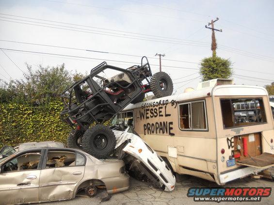 Off-road buggy on top of a beige bus, with wrecked cars underneath. 'Wessel Propane' is written on the bus