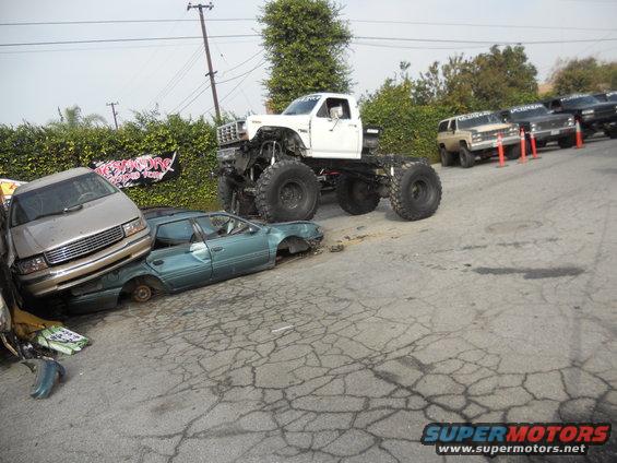White monster truck over crushed cars, with other trucks in background