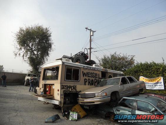 Modified RV with cars stacked on top and in front. Graffiti on RV reads 'WESSEL PROPA' and 'DESMADRE'