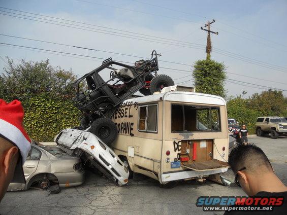Several wrecked cars are piled up, with a bus on top of some of them. A buggy is on the bus roof. Two people are watching