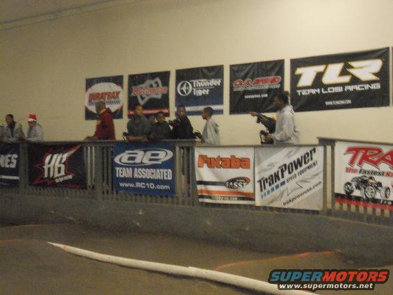 People watch an RC car race from a spectator area with sponsor banners on the wall