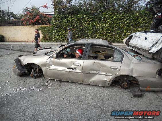 Damaged sedan with wheels missing, person in Santa hat in driver's seat. Another wrecked vehicle is on top