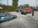 1986 Ford Bronco - A collection of modified trucks and a heavily damaged sedan are parked in a lot, suggesting a recent event or gathering.