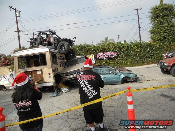 Scene of multiple wrecked vehicles, including a bus with a buggy on top, and people wearing Santa hats
