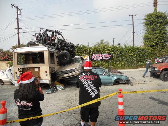Several wrecked cars and a bus are piled up, with a buggy on top of the bus. People in Santa hats are present