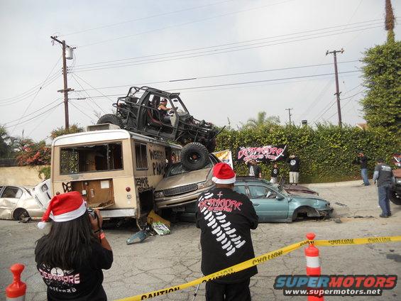 Crushed car under bus with roll cage vehicle on top, people in Santa hats watching